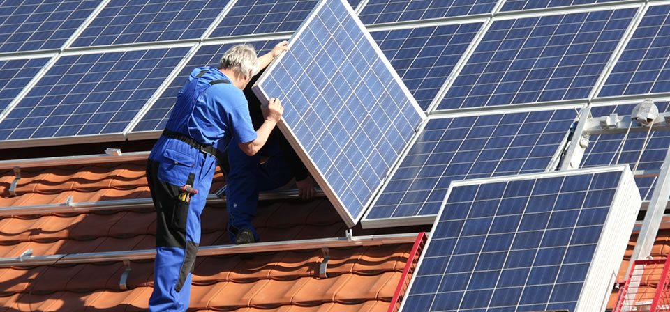 Handwerker in blauer Arbeitskleidung installieren Solarmodule auf einem roten Ziegeldach. Elektro Bohr in Saalfeld.